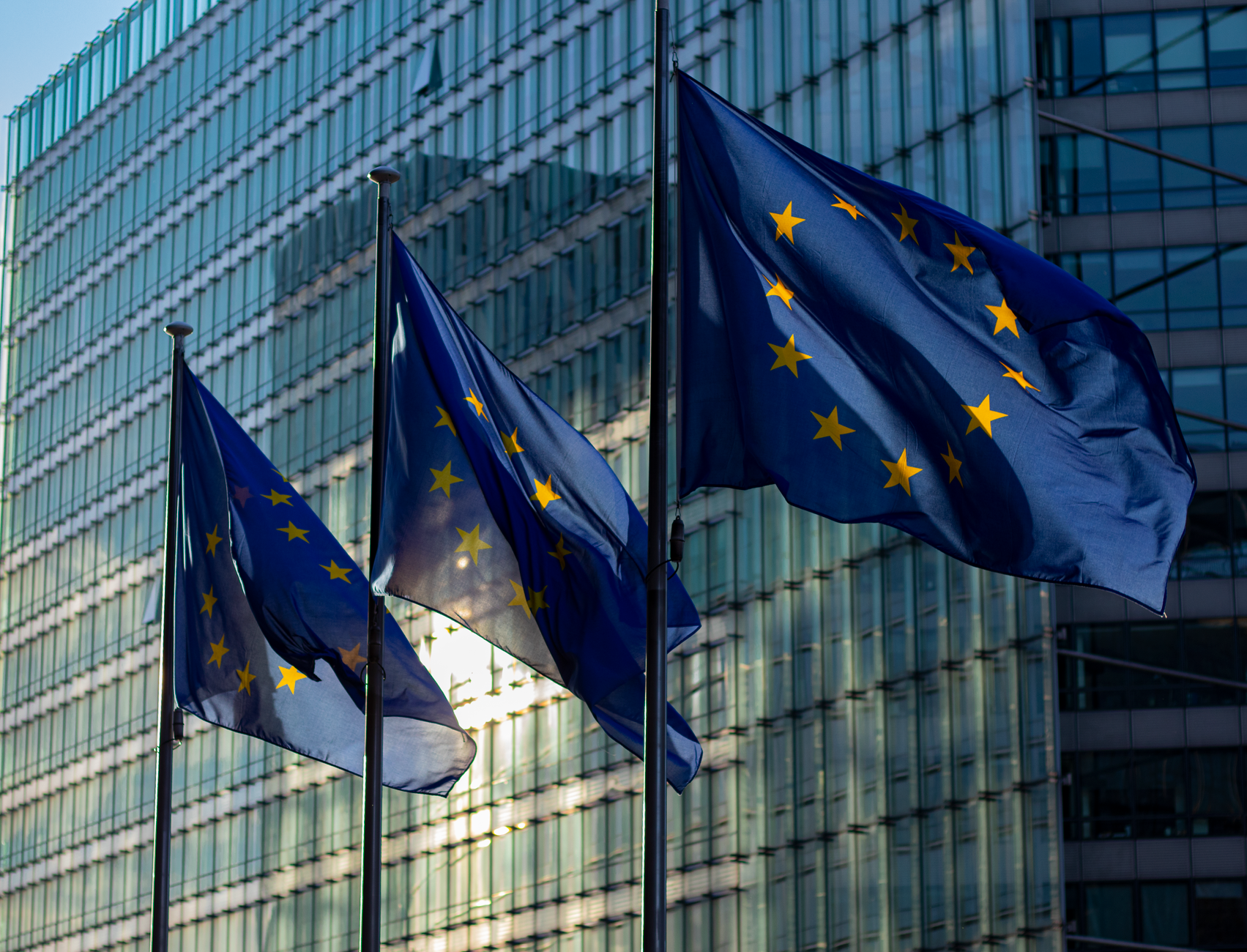 Drei Fahnen der Europäischen Union vor der Glasfassade eines Verwaltungsgebäudes in Brüssel.
