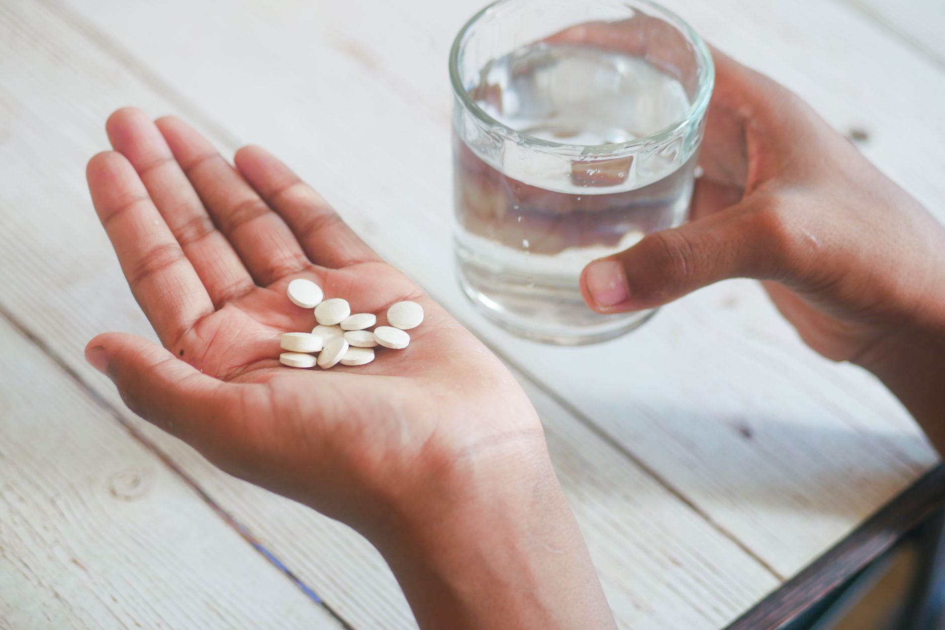 Hand mit mehreren weißen Tabletten neben einem Glas Wasser