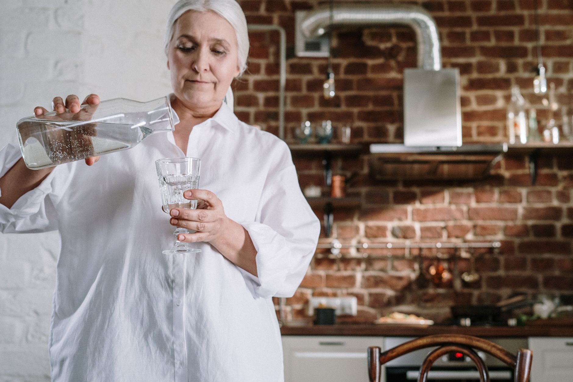 Una donna anziana in camicia bianca versa acqua da una caraffa di vetro in un bicchiere, in cucina