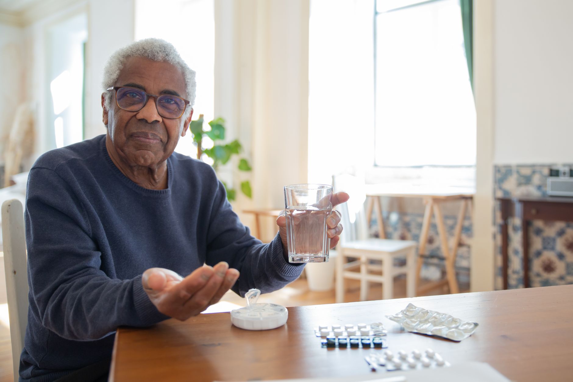 Homme âgé assis à une table en bois, tenant un verre d'eau, des plaquettes de comprimés posées devant lui.