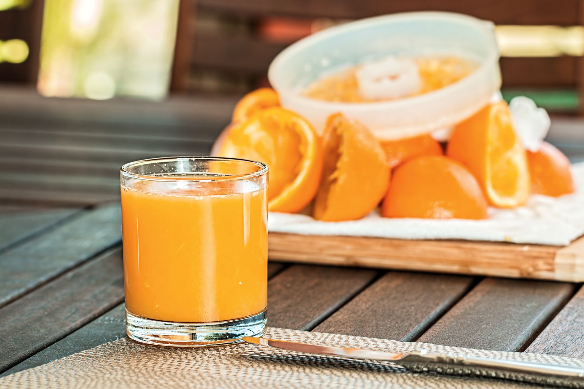 Glas frisch gepresster Orangensaft auf einem Holztisch, dahinter ausgepresste Orangenhälften und eine Saftpresse.