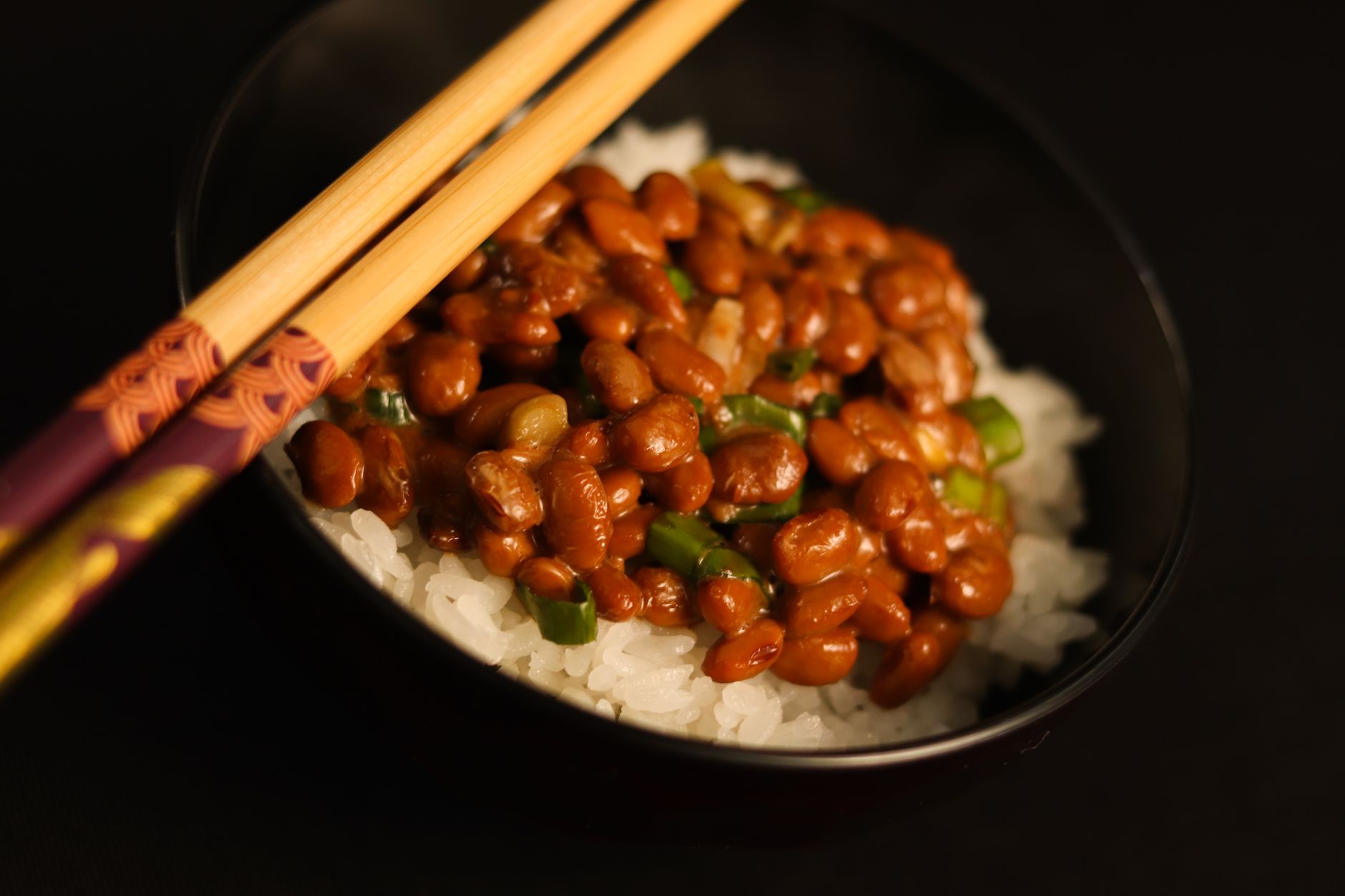 Schale mit Natto und Reis, daneben Essstäbchen, auf dunklem Untergrund.