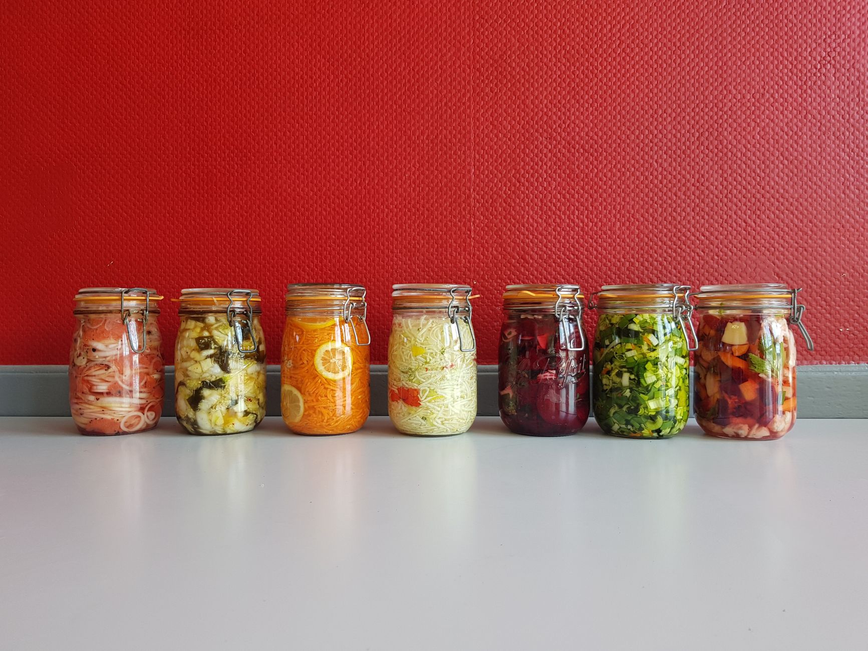 Sieben Bügelgläser mit fermentiertem Gemüse vor roter Wand