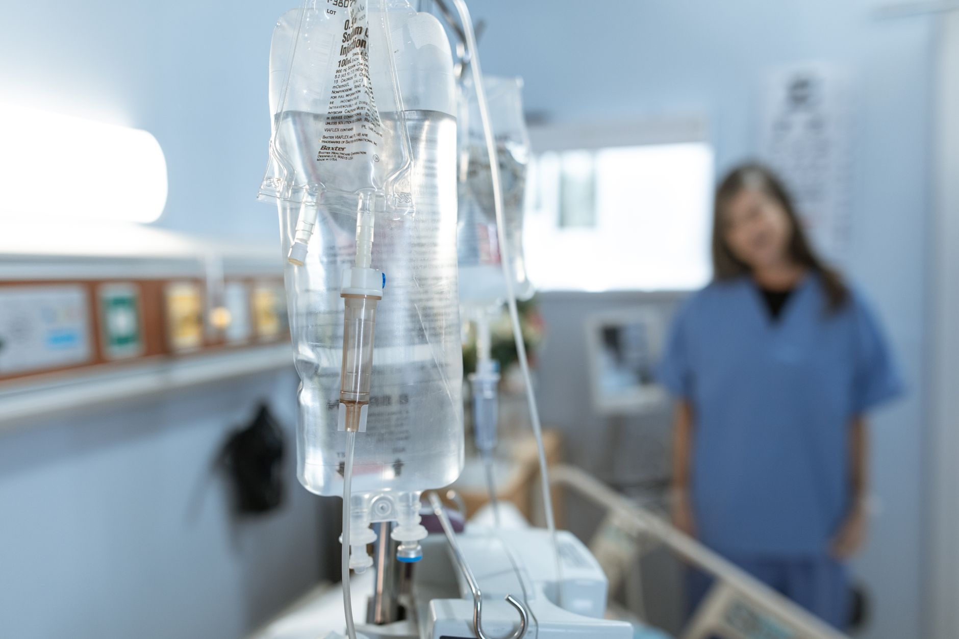 Infusionsbeutel an einem Ständer, im Hintergrund unscharf eine Pflegekraft.