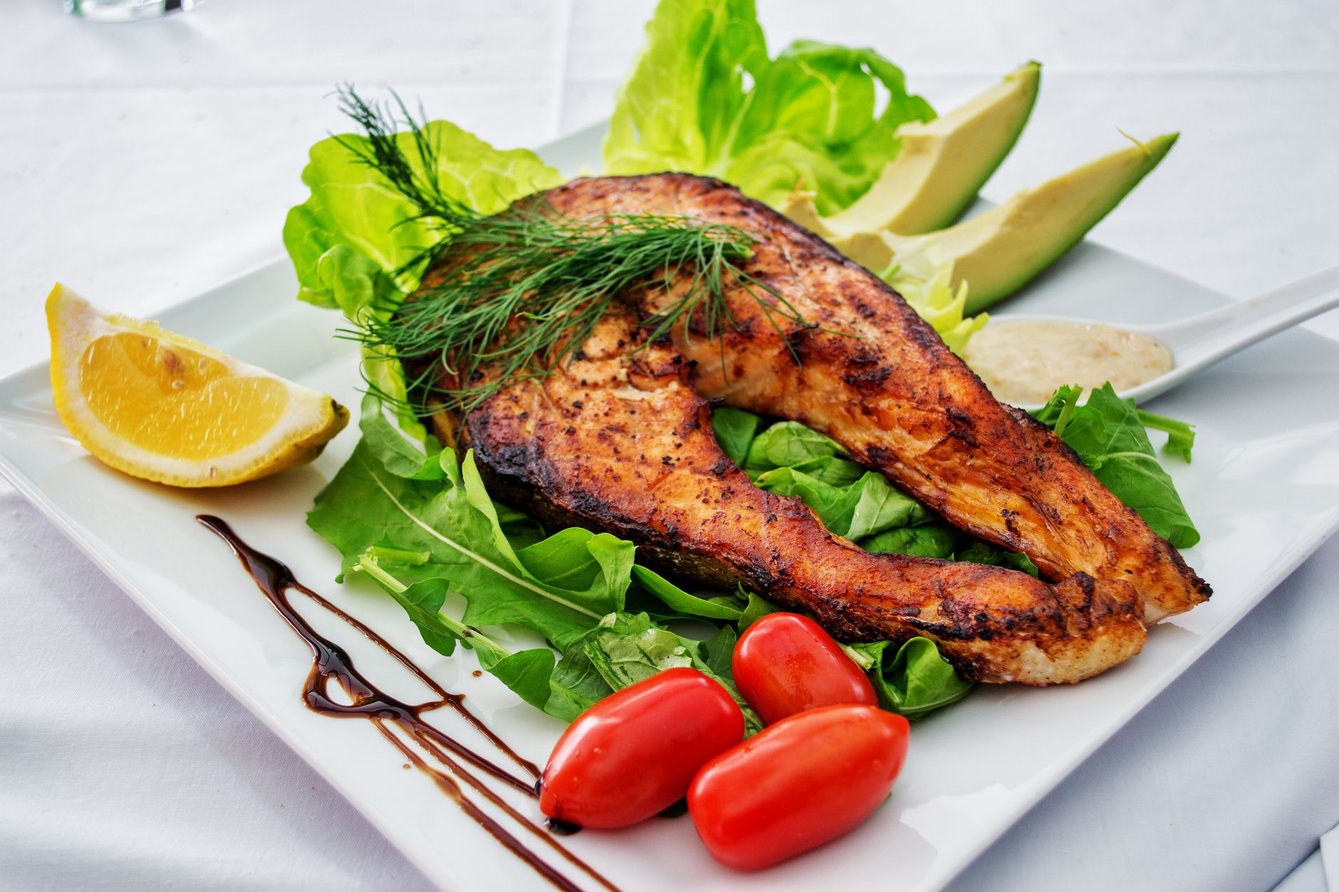Gegrilltes Lachssteak mit Avocado, Rucola und Zitrone auf einem weißen Teller.