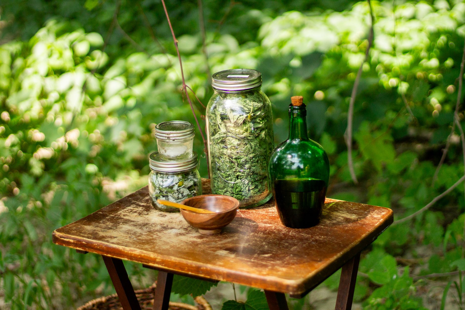 Gläser mit getrockneten Kräutern und eine dunkle Flasche auf einem Holztisch im Grünen