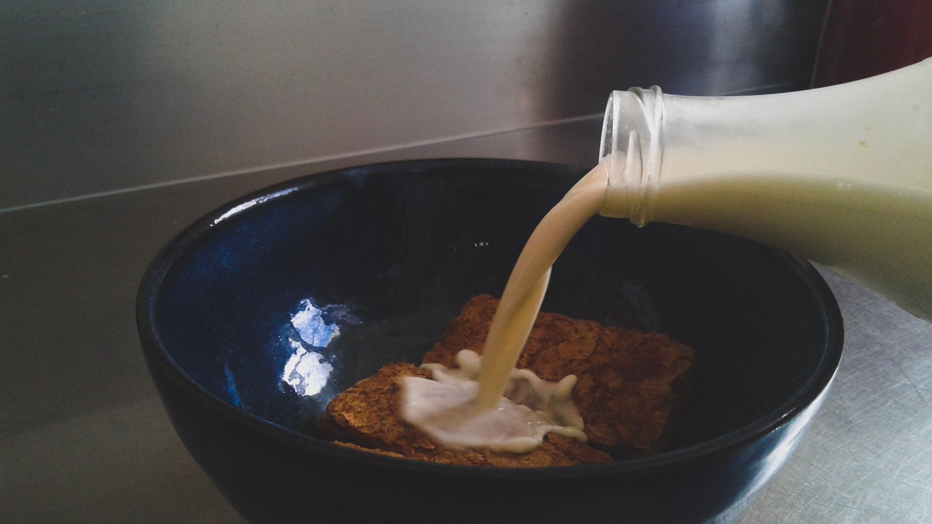 Milch wird aus einer Flasche über Vollkornflocken in eine dunkle Schale gegossen